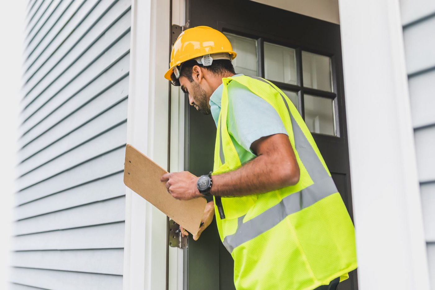 Inspector in a hard hat and safety vest examining a door frame while taking notes on a clipboard